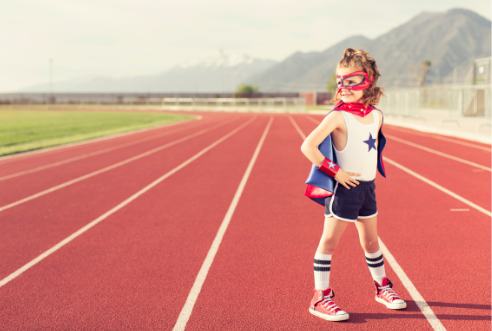A child dressed as a superhero stands confidently on a running track, ready to race.